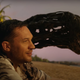Eddie Brock, in a Hawaiian shirt, sits happily on a beach while talking to the distended head of Venom.